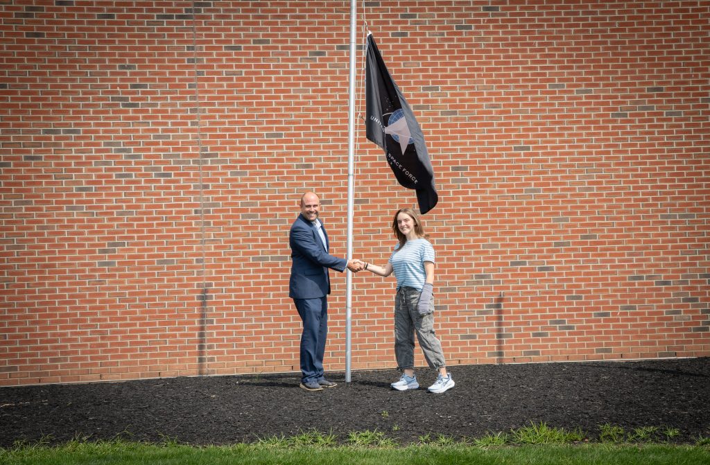 Gia shakes hands with Secondary Principal Richard Cummings in front of the installed Space Force flag outside the school.